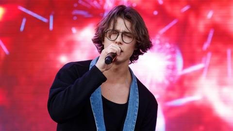 David Kushner singing into a microphone with his eyes closed wearing a black jacket with a denim collar 