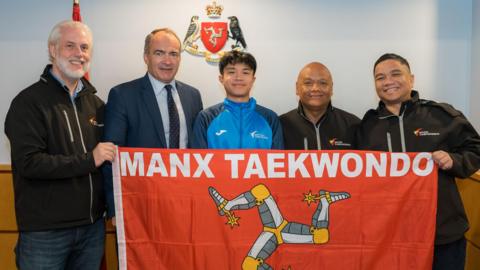 Men in sports tracksuits, along with the Chief Minister in a suit and 18-year-old Nino Ontoy wearing a blue tracksuit, they hold a Manx flag which reads Manx Taekwondo.