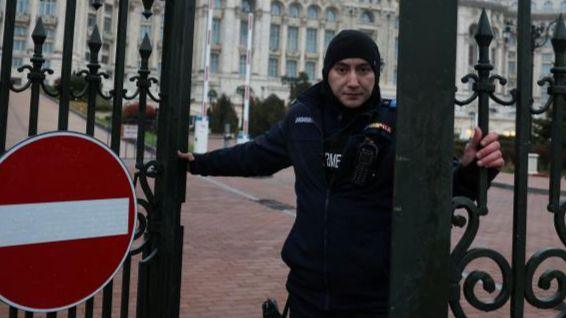 A police officer, wearing a dark hood and jacket, closes a wrought iron gate bearing a no entry sign at the grand Palace of Parliament