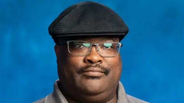 JoJo Burgess stands in front of a blue background. He sports a mustache, glasses and a black hat