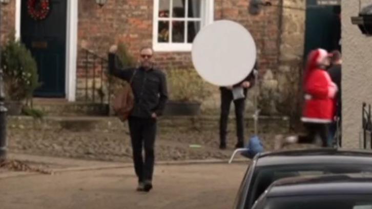 Mr Sutherland walks down a path with a brown bag slung over his shoulder. He is wearing sunglasses and is dressed in dark clothes, with one hand in his pocket and the other raised in a thumbs-up. A microphone can be seen in the right corner.