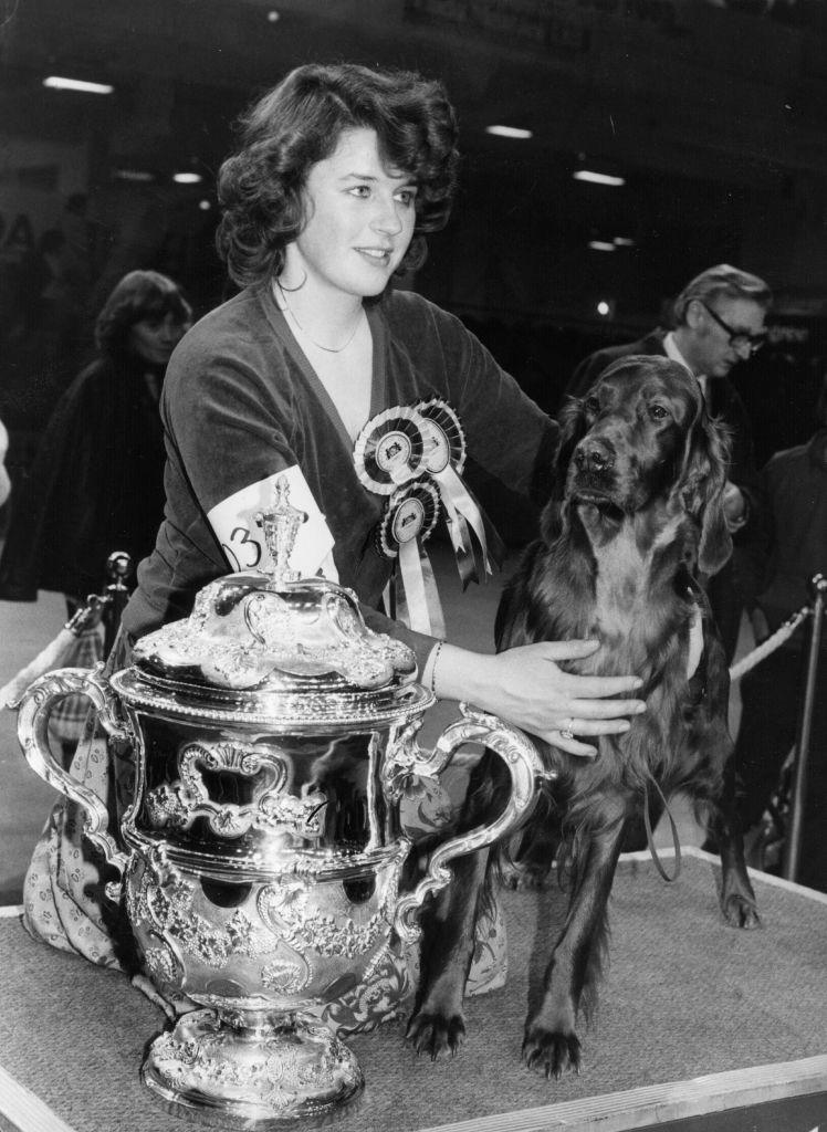 A woman with short dark hair is wearing a black top and holding her dog on a table. There is a large trophy sitting on the table. The woman is wearing lots of medals.