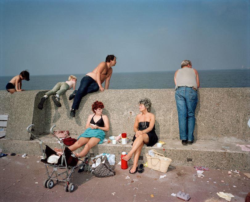 A family sitting on a stone wall with the sea behind them, they are enjoying a picnic and there is litter surrounding them on the floor 