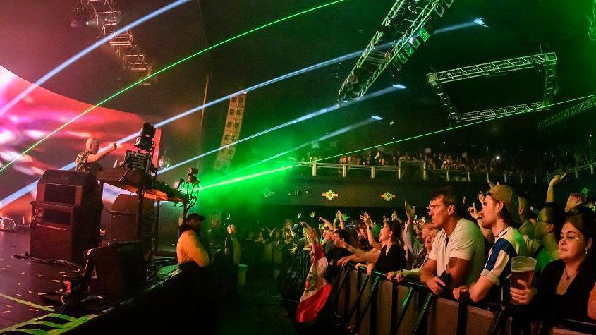 People at a front row of a concert, right, watch Fatboy Slim on stage behind a DJ set of decks, left