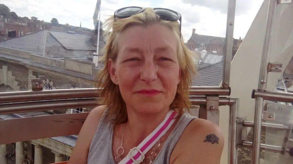 Dawn Sturgess has short blonde hair and dark sunglasses on her head, She is wearing a grey vest top, silver chain necklace and the pink and white strap of bag is crossing her body. She is sitting on a balcony with rooftops visible in the background