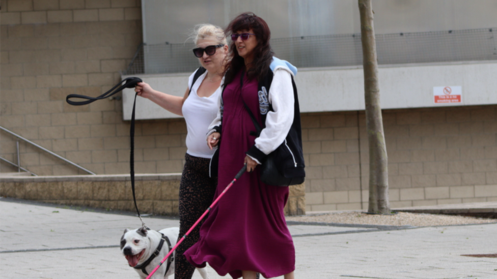 Photograph of two women walking arm in arm along the pavement. The woman on the left is blonde and holds the lead for a white dog and the woman on the right has brown hair, wears a magenta dress and is holding a pink cane. They both wear sunglasses.