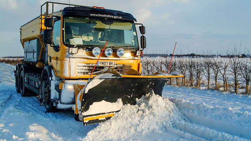 A bright yellow Lincolnshire County Council gritting lorry drives along a country lane pushing snow out of the way with a snow plough attached to the front