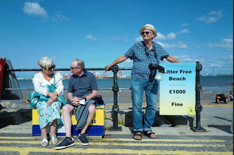 An older man wearing sunglasses, a summer shirt, hat and jeans and sandals, leans on a black fence on a promenade on a sunny day, next to an older couple. A camera is hanging around his neck 