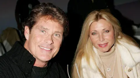 Getty Images Hasselhoff and Bach seen seated beside each other in 2005. He wears a black sweater and she has long blonde hair, is wearing sparkly jewelry and a white coat.