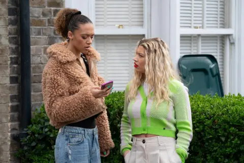 BBC/Jack Barnes/Kieron McCarron Francesca Henry playing Gina Knight (left) holds her phone next to and Molly Rainford, playing Anna Knight (right) in EastEnders. A woman with brown hair and a fluffy coat holds her phone standing next to a woman with blonde hair and a green jumper.