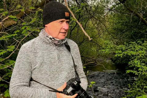 Tom Kelly Tom is wearing a black woolly hat and grey jumper. He is holding a camera with a long zoom lens and is standing beside a river with tree branches over head.