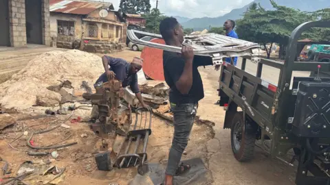 Scrap metal dealers at work by the side of a road in Morogoro city centre. A man holds the shell of a car door as he moves to place in the back of a lorry. Someone else is behind him holding two pieces of metal on the ground.