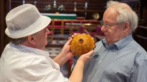 MARTIN ELLIOTT Mike Holling is wearing a white uniform and white hat. He is holding a pie up to the face of Matthew who is inspecting it. Matthew is wearing glasses and has white hair.