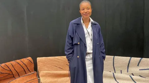 Adaobi Tricia Nwaubani Kelechi Ejikeme in a long navy coat standing in front of jute rugs - mainly brown and beige with navy lined designs on them - at the Decorex show in London