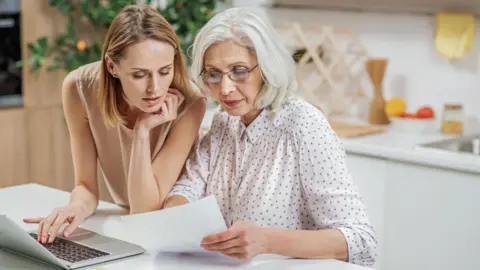 Getty Images A young woman looks at paperwork with a woman who is a generation older. A laptop is open in front of them.