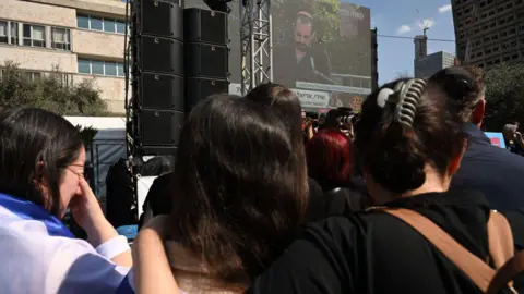 Getty Images Israelis watch a big screen showing Yarden Bibas speaking at the funeral of his wife Shiri and sons Ariel and Kfir, in Tel Aviv, Israel (26 February 2025)