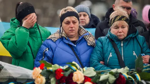 BBC/Matthew Goddard Natalya is flanked by two other women at her husband's funeral, with flowers visible in the foreground.