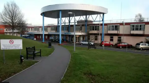 PA Media View of two-storey Hinchingbrooke Hospital, showing lawn, a pathway, a line of parked cars and a covered area over the main entrance 