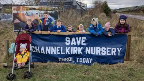 A group of children of various ages line up behind a sign reading Save Channelkirk Nursery. One child sits in a pushchair in front of the sign