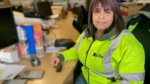 Heidi Tomlinson/BBC A woman in a hi-vis jacket sits at a desk 