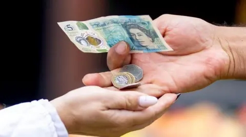 Getty Images Two hands with a £5 note and coins being passed over.