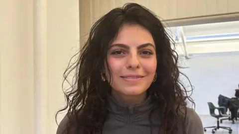 BBC Sarah Dattani Tucker leans forwards and smiles at the camera. She has long curly dark hair worn loose and wears a zip-up grey top. She's pictured in a well lit white-painted room. 