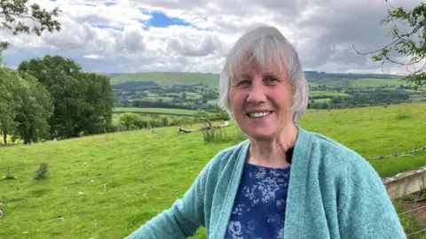 BBC Dina Smith in turquoise cardigan leaning on a fence in green valleys