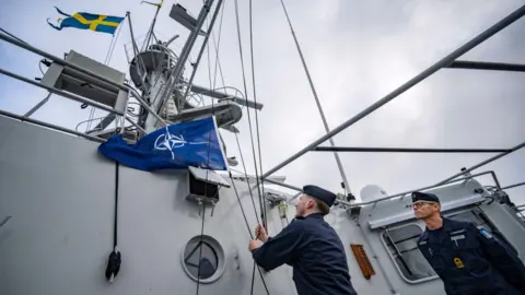 Getty Images Two sailors on a military vessel. One of them is raising the NATO flag next to flag of Sweden while the other looks on. 