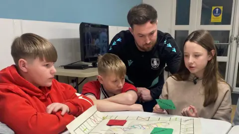 Two boys and a girl at Wincobank youth club look at a boardgame they've designed, with an adult also looking at the game