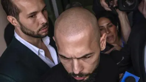 Man with a shaved head looking down as he is jostled by reporters and cameramen. To his left is another man wearing a white open collar shirt and a dark blue suit