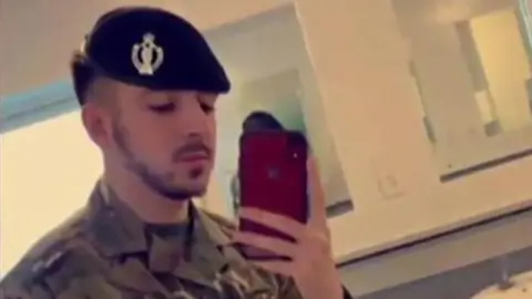 Selfie of Kyle Clifford wearing his army uniform and beret in a bathroom. He is holding a red phone and has a dark, close-cropped beard and moustache.