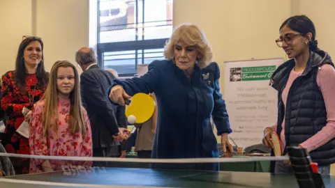 Reuters Queen Camilla playing table tennis with a yellow paddle. She is wearing a blue coat and a broach. Next to her are two children watching as well as a woman in a red dress.