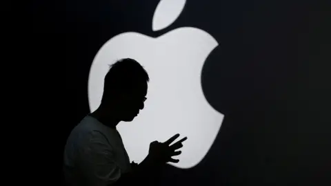 Silhouette of a man holding a smartphone in front of a lit-up Apple logo.