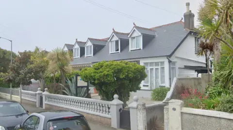 A white house, with windows in the eaves and palm trees in the front garden.