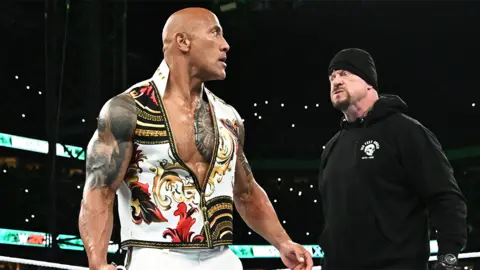 The Rock, a muscular man with tattoos looking behind him to The Undertaker, a man wearing all black.