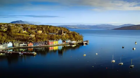 An aerial view of Tobermory, and its brightly coloured houses, and its bay with yachts anchored in it. There are hills on the coast in the background.