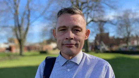 BBC A man with short grey hair and wearing a blue patterned shirt with a piercing above his left eye. He is standing in a field and looking into the camera.