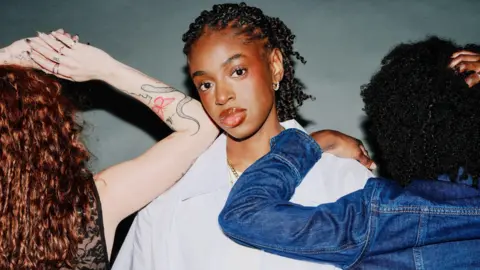 Reuben Bastienne Cat Burns in a promotional shot for her new single. Cat wears an oversized white shirt, gold chain, and has her braided hair tied back. Two women, facing the opposite direction, stand on either side of her. On the left, a red-headed woman with long curly hair rests her tattooed arm on Cat's shoulder while on her right, a woman with curly dark hair wearing a denim jacket rests her hand at the top of her back. 