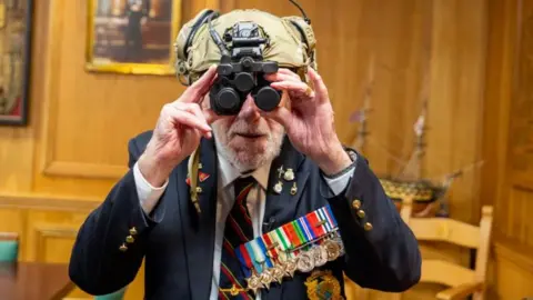 Royal Navy Mr Eskdale wearing a blue suit with a white shirt and navy blue and red tie. He is also wearing several medals while he wears a modern military helmet with night vision. 