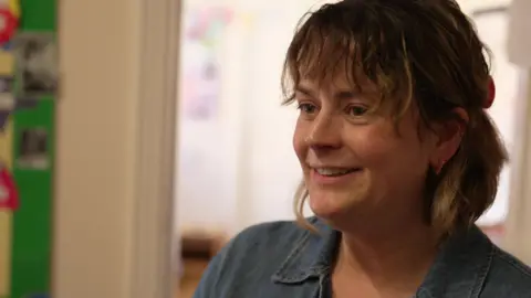 Lady with short dark blonde hair and fringe, wearing denim shirt, smiling at the camera. We can only see her face and shoulders