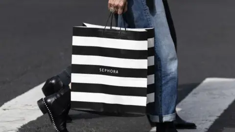 A woman is wearing blue jeans and black boots. She is holding a black and white striped Sephora bag and is walking across a zebra crossing