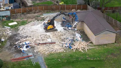 PA Media A digger demolishes the spa pool block at the home of Hannah Ingram-Moore, the daughter of the late Captain Sir Tom Moore, at Marston Moretaine, Bedfordshire. The picture is taken from above, and debris is spread across the grass