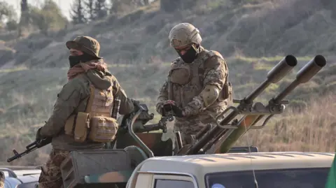 Two Syrian security force soldiers sit on top of a pick up truck armed with a  twin-barrelled heavy machine gun. Both men have masks on their faces and are dressed in full military fighting equipment and camoflage uniforms.