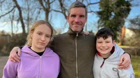 Neil Duckett, wearing a dark green zip-up fleece, stood between his daughter Toula, wearing a purple hoodie, and Joe who is wearing a light grey hoodie. Neil has his arms around his children's shoulders.