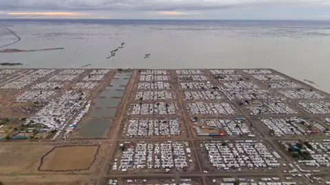 Drone image of part of rectangular camp surrounded by water and covered in tightly-packed huts