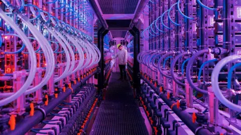 Vaxa A walkway lined with bioreactors giving off pink and purple light. At one end a worker examines the equipment.