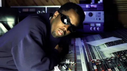 Getty Images Sean "Diddy" Combs, wearing sunglasses and a baggy navy sweatshirt, with an stud in his right ear, neatly trimmed hair and a goatee, leaning over the mixing desk of his recording studio, which is strewn with papers and Sharpie pens