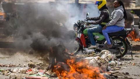 A man and woman on a motorbike driving past a pile of burning rubbish on the road