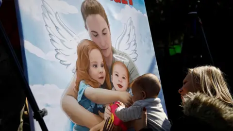 Reuters A woman holds up a baby next to artwork depicting the dead Israeli hostages Shiri, Ariel and Kfir Bibas, in Tel Aviv, Israel, during their funeral procession (26 February 2025)