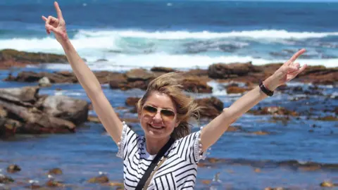 Nathan Angell Allison pictured with her arms in the air smiling in the sunshine. She wears glasses and has a navy and white striped top on. 